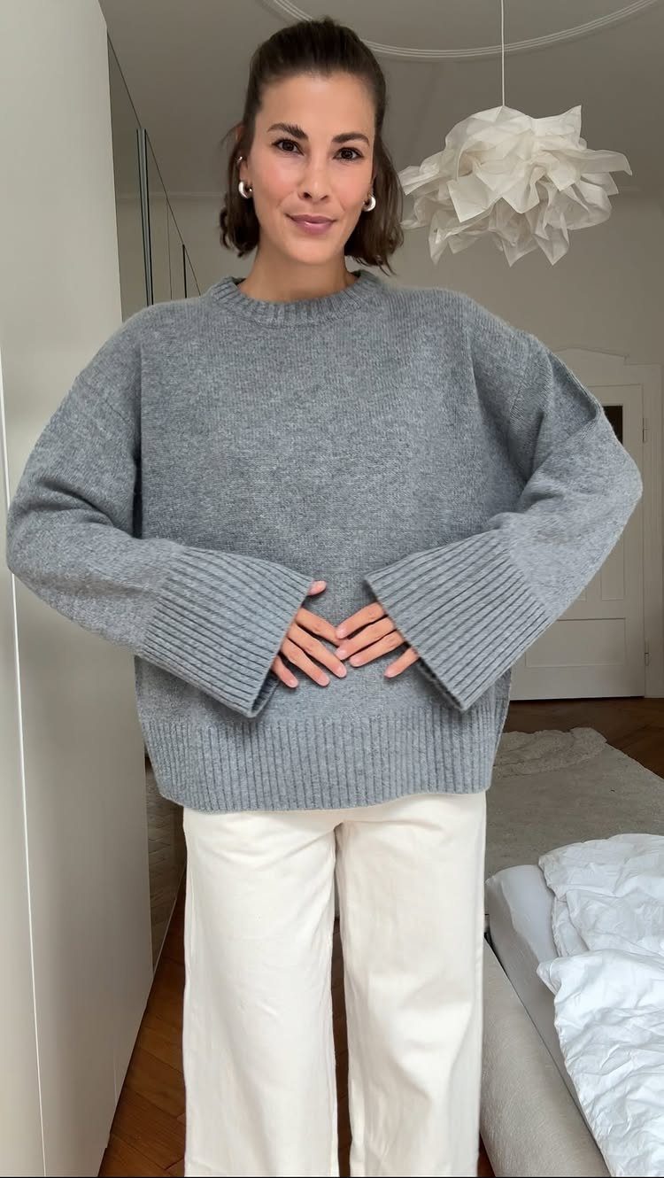 A woman wearing a grey oversized sweater and white trousers standing in a bright room.