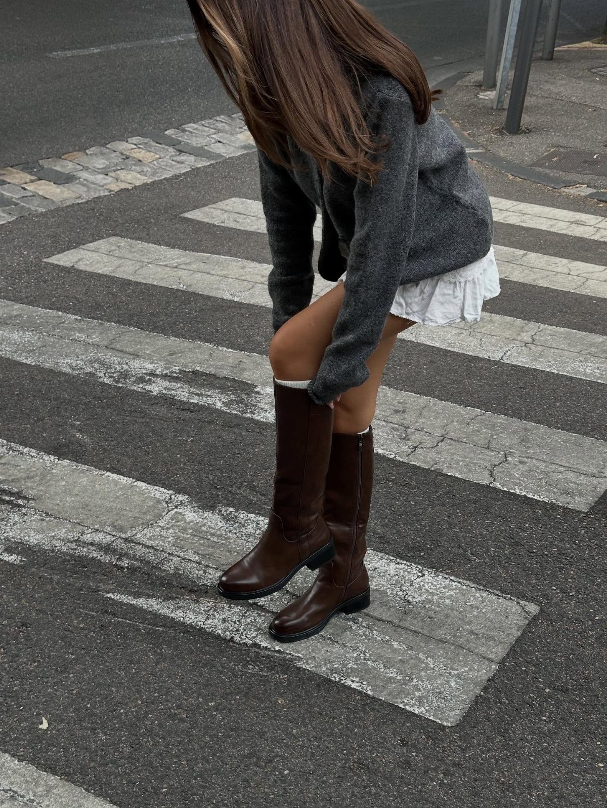 A person wearing a grey sweater, white skirt, and tall brown boots on a crosswalk.