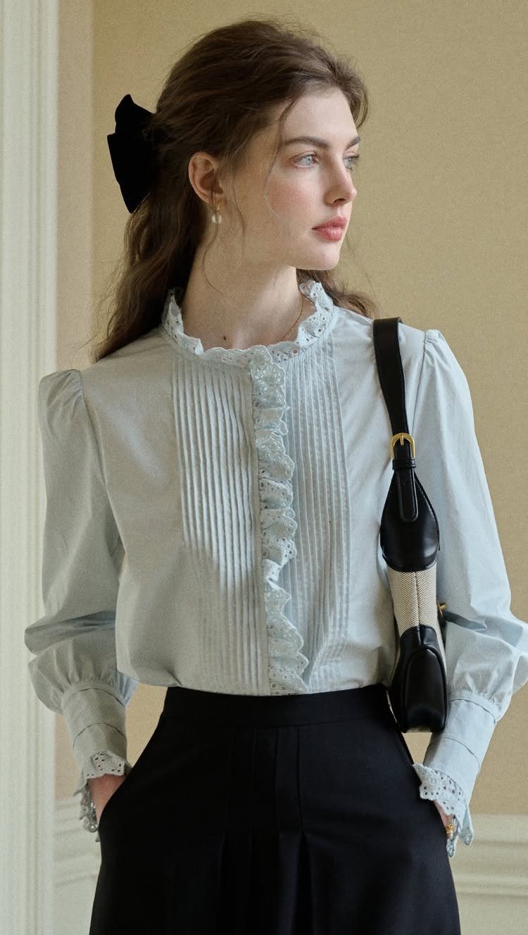 A young woman wearing a light blue ruffled blouse, black trousers, and a black hair bow.