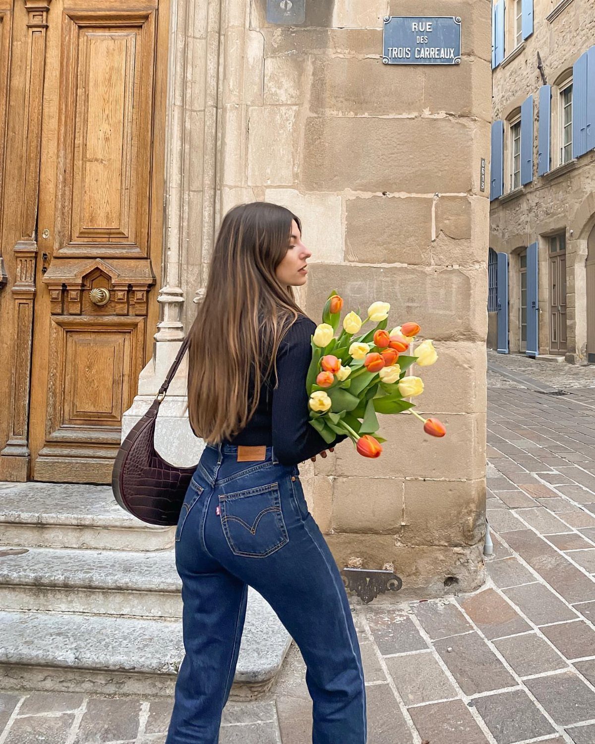 A woman in dark blue straight-leg jeans and a black top carries yellow tulips outdoors.