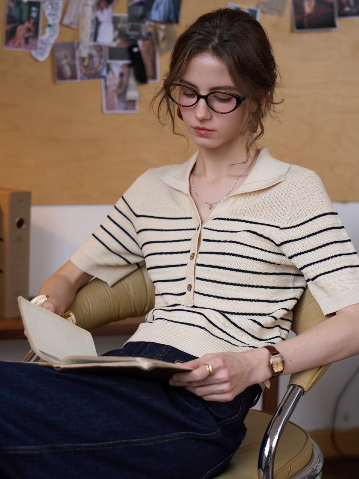 A woman wearing a striped polo shirt sits reading an open notebook in a room.