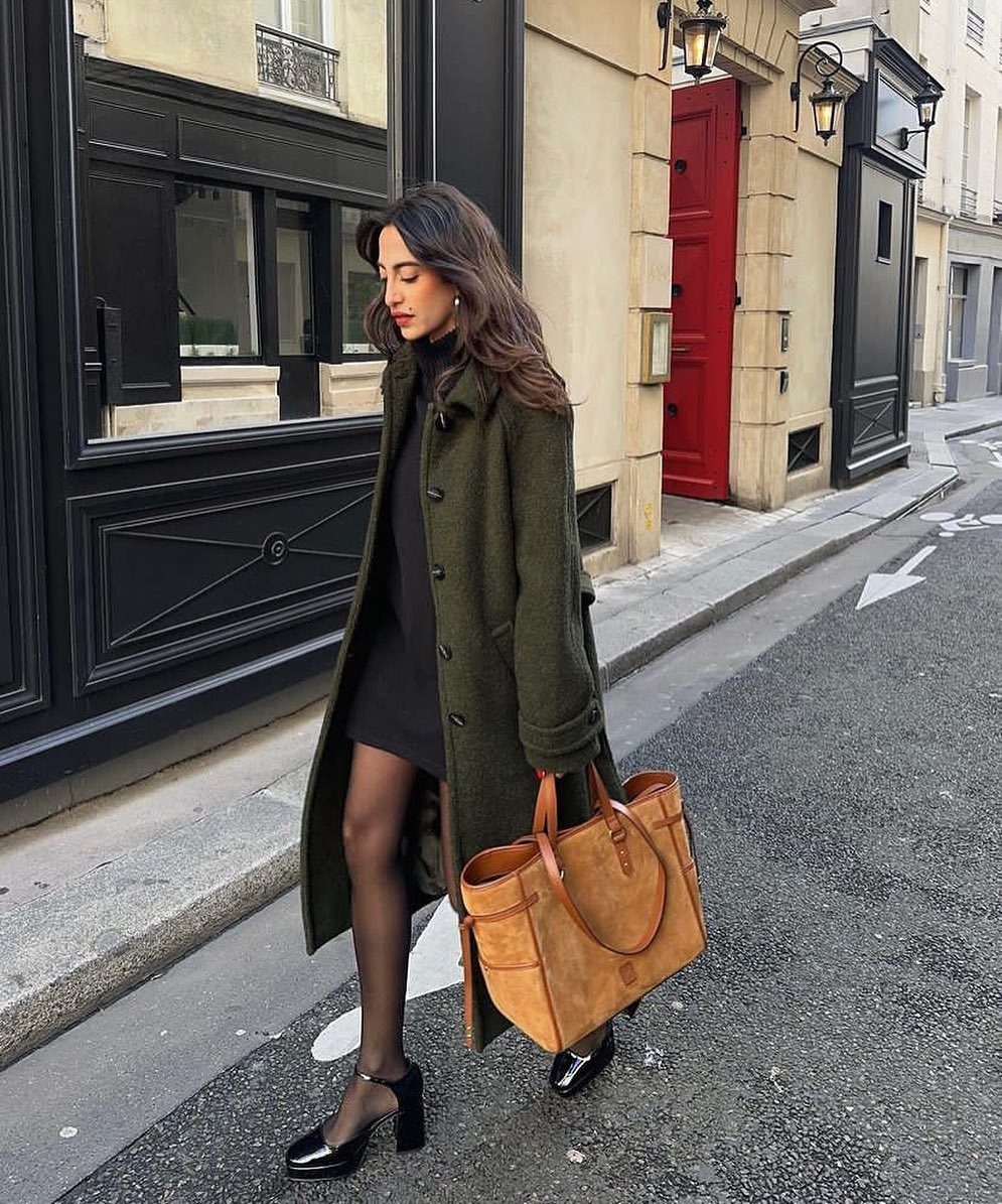A woman in an olive coat walking on a city street carrying a tan leather tote bag.