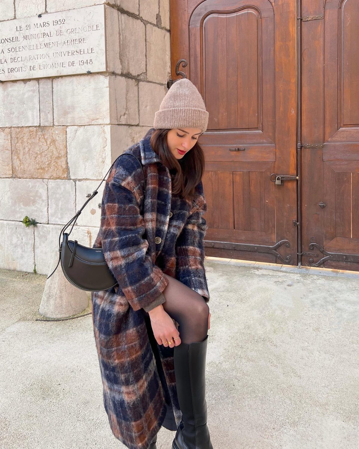 Woman in a plaid wool coat and beige beanie adjusting her tall black boots outdoors.
