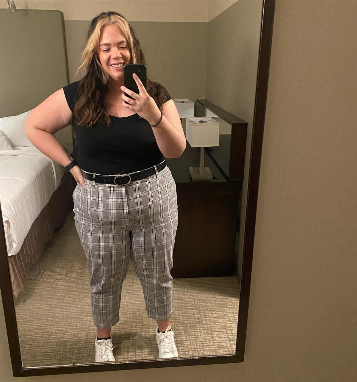 A smiling woman in a black top and plaid trousers taking a mirror selfie.