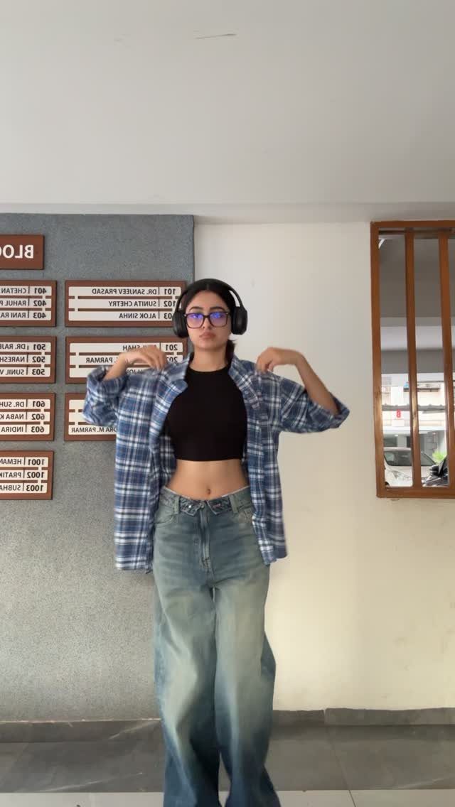 A young woman posing in an open blue plaid flannel shirt and wide-leg denim jeans.