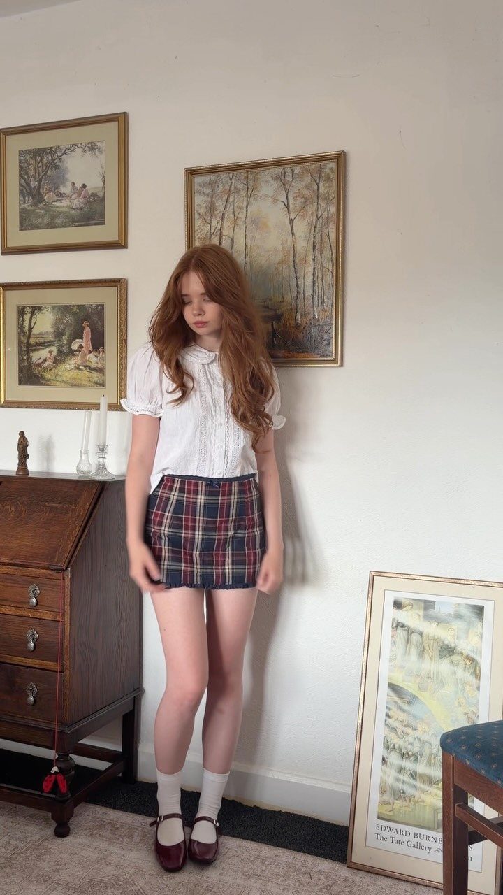 A young woman with red hair wearing a white blouse, plaid skirt, and dark Mary Jane shoes.