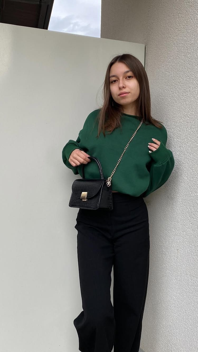 A young woman posing in a dark green sweater and black trousers with a small handbag.