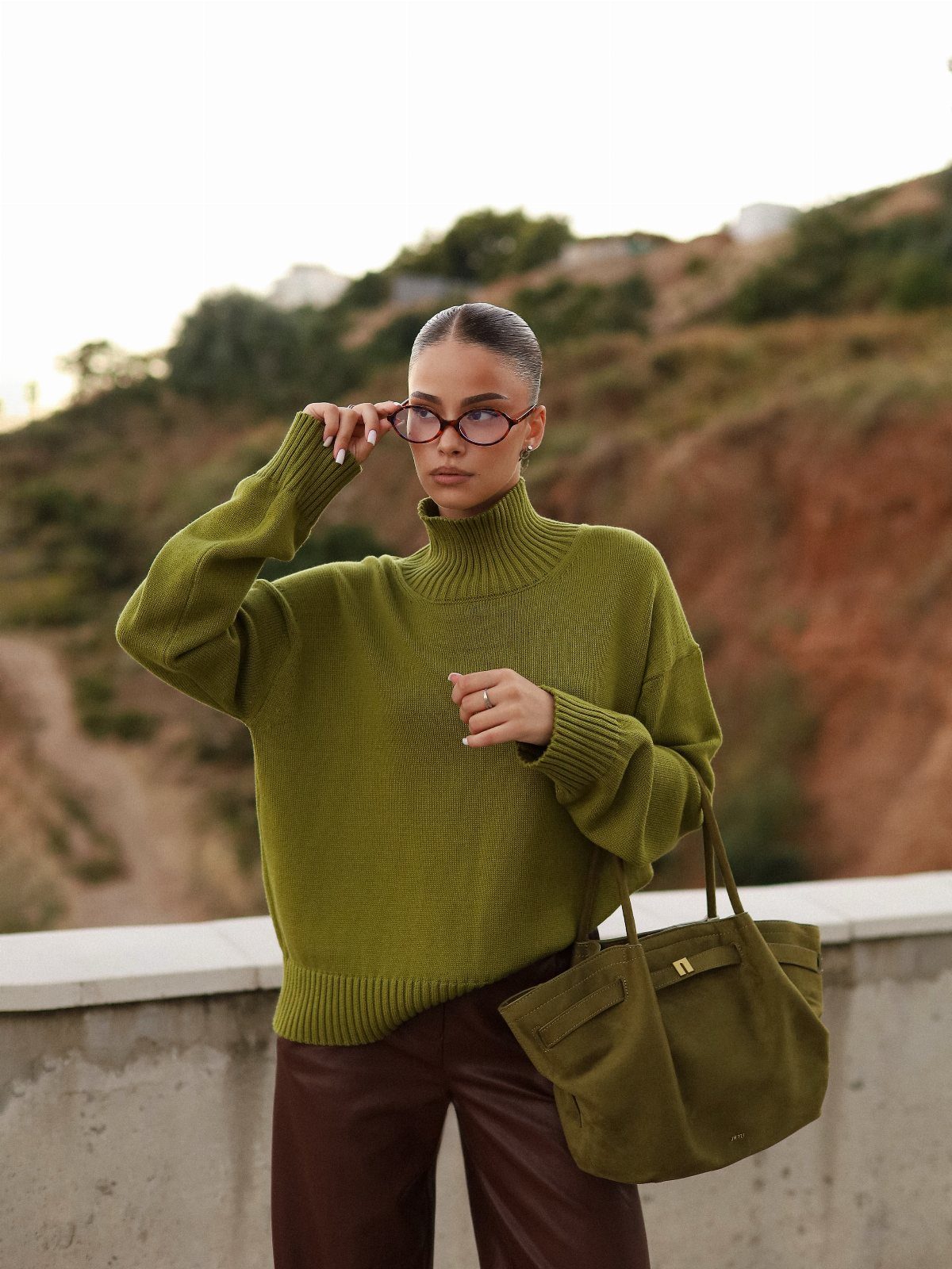 A woman wearing an olive green turtleneck sweater and brown leather pants outdoors.
