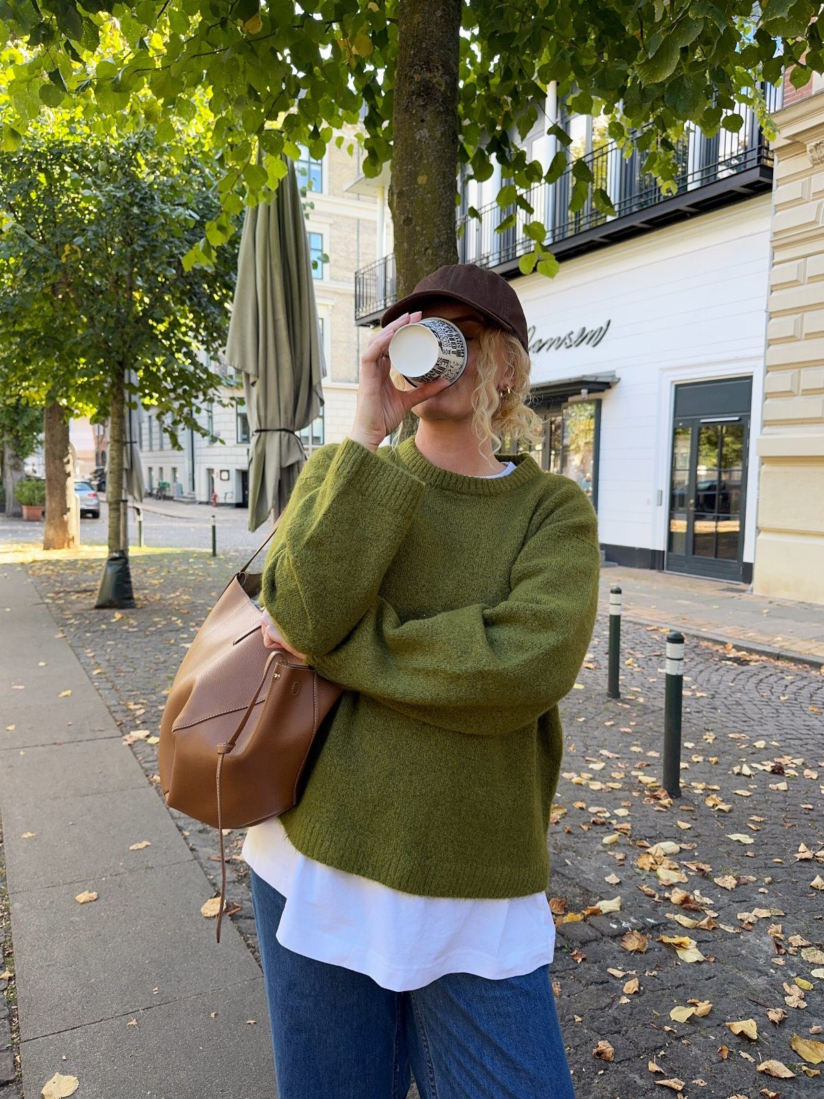 A person wearing an olive green sweater and blue jeans holding a coffee cup outdoors.