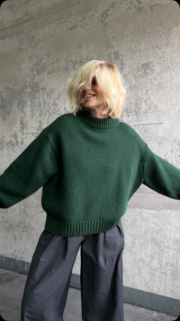 Blonde woman wearing a dark green oversized sweater and wide-leg grey trousers against a concrete wall.