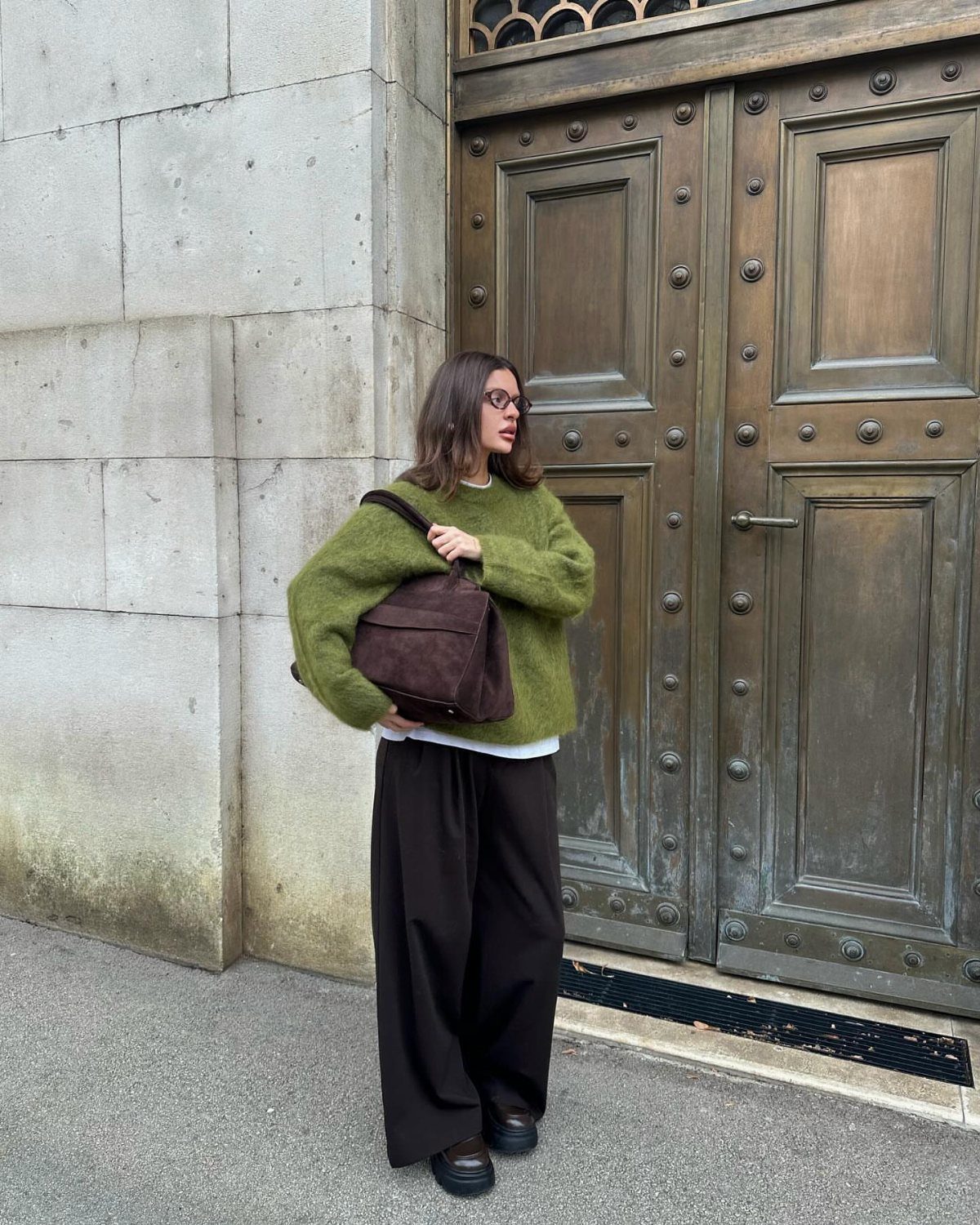 Woman in a fuzzy green sweater and wide-leg black trousers standing by large bronze doors.