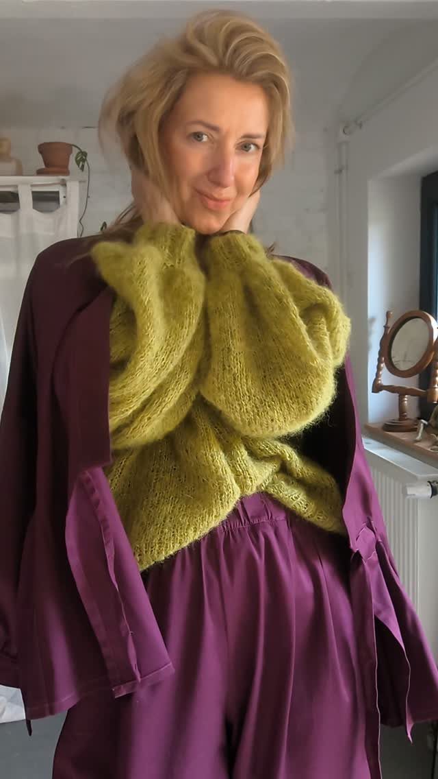 Woman posing in a lime green sweater and flowing purple satin trousers indoors.