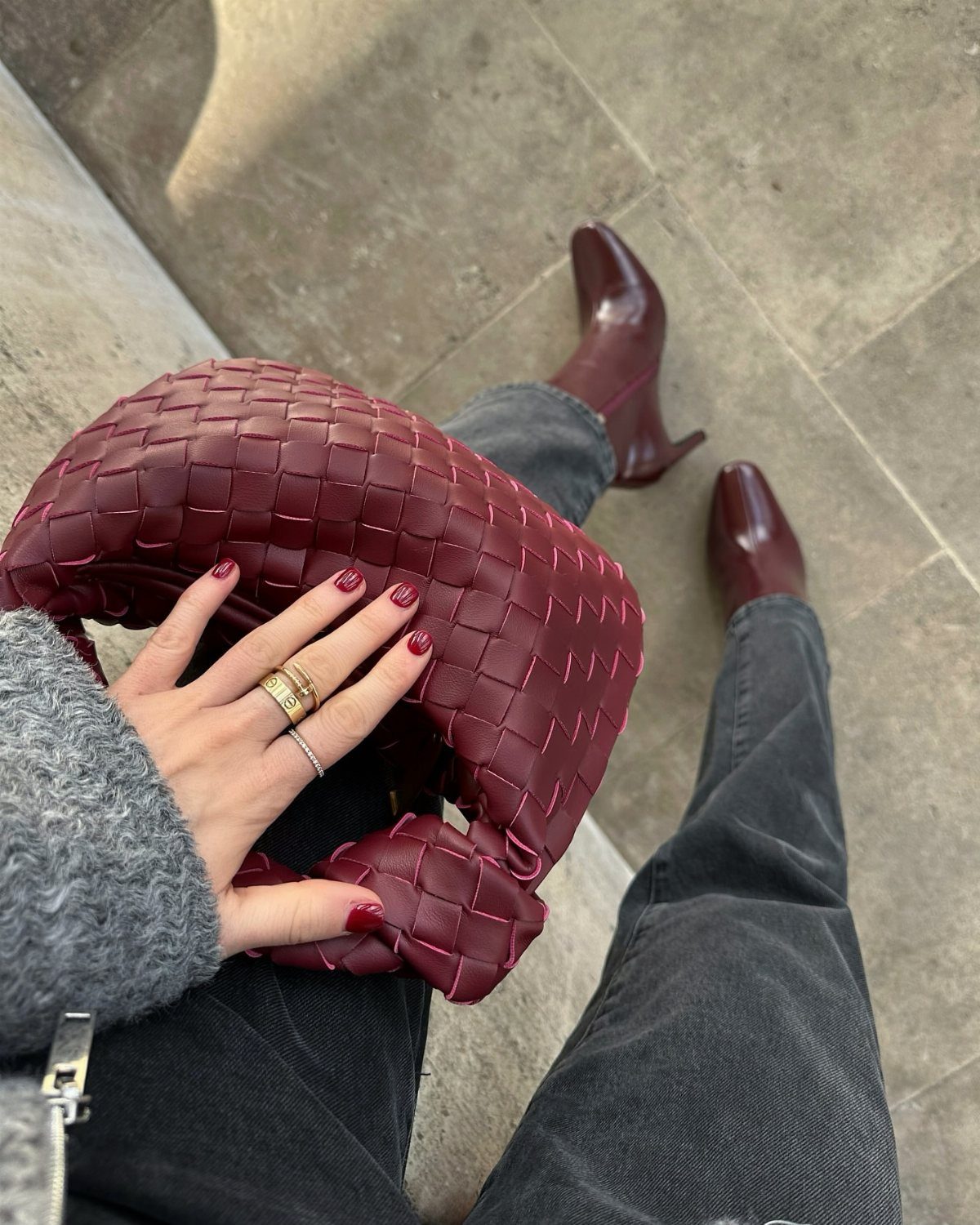 Person wearing grey jeans with burgundy boots, a burgundy woven handbag, and burgundy nail polish.