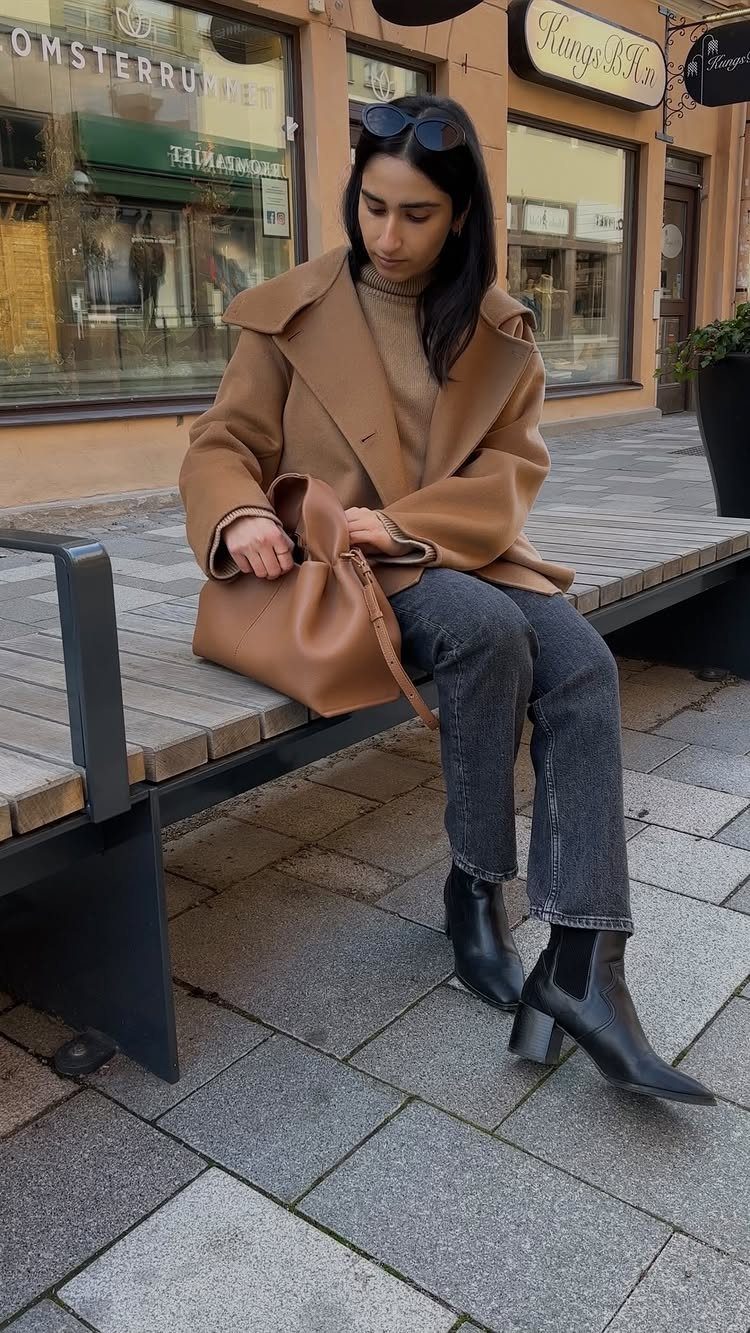 Woman wearing grey jeans, a tan turtleneck, and a camel coat sitting on a bench.
