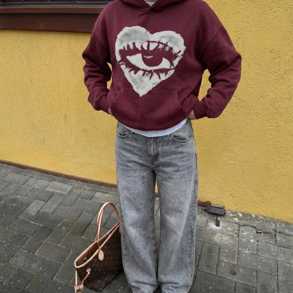 Person wearing a maroon graphic hoodie and grey jeans standing against a yellow wall.