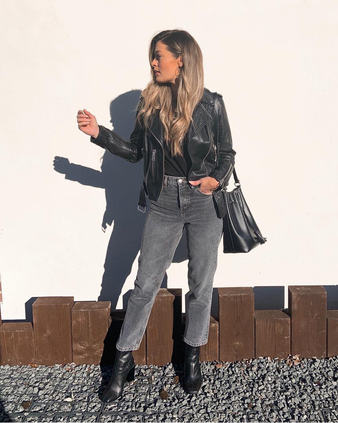A woman wearing grey jeans and a black leather jacket standing against a white wall.