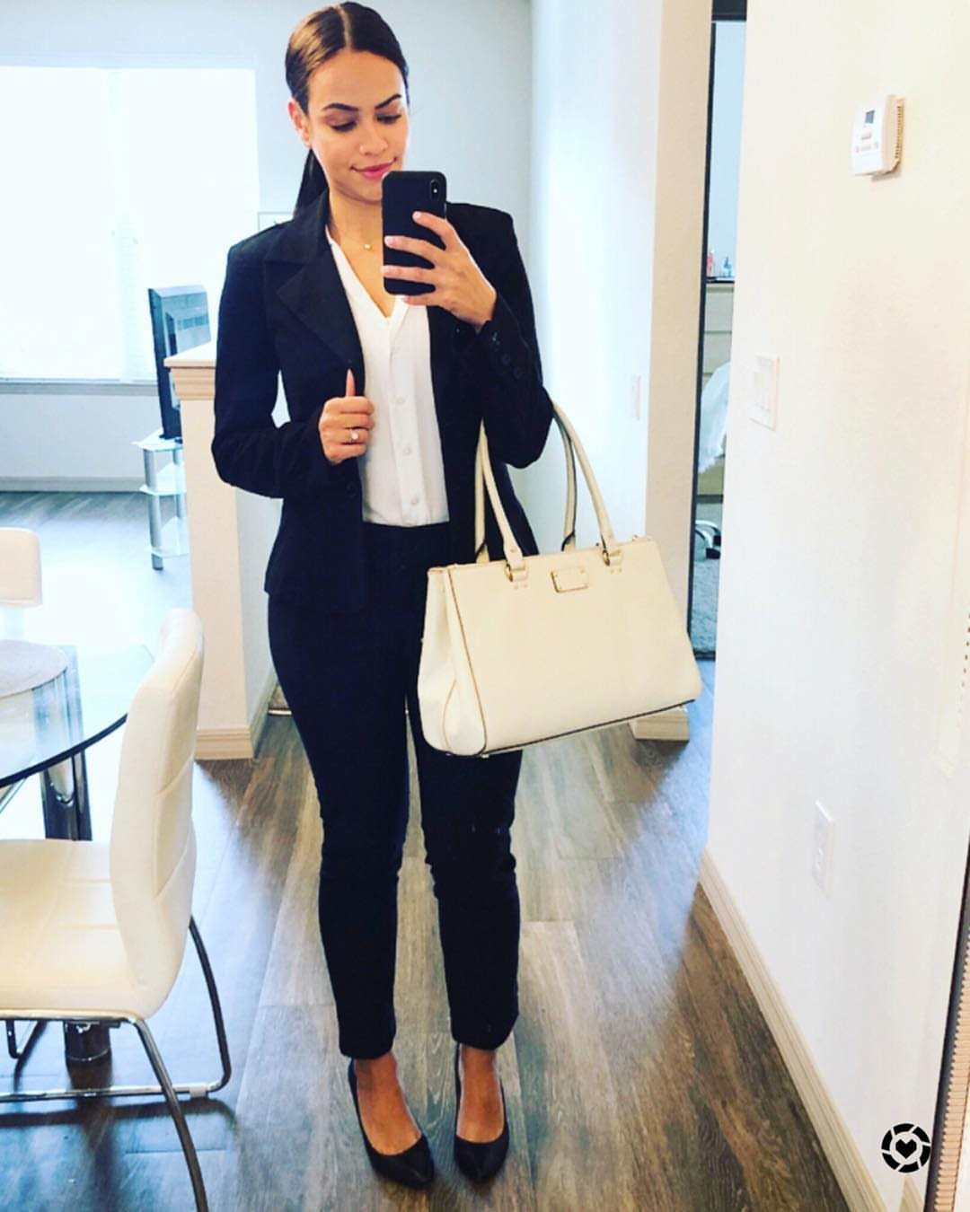 A woman in a black pantsuit and white blouse posing for a mirror selfie.