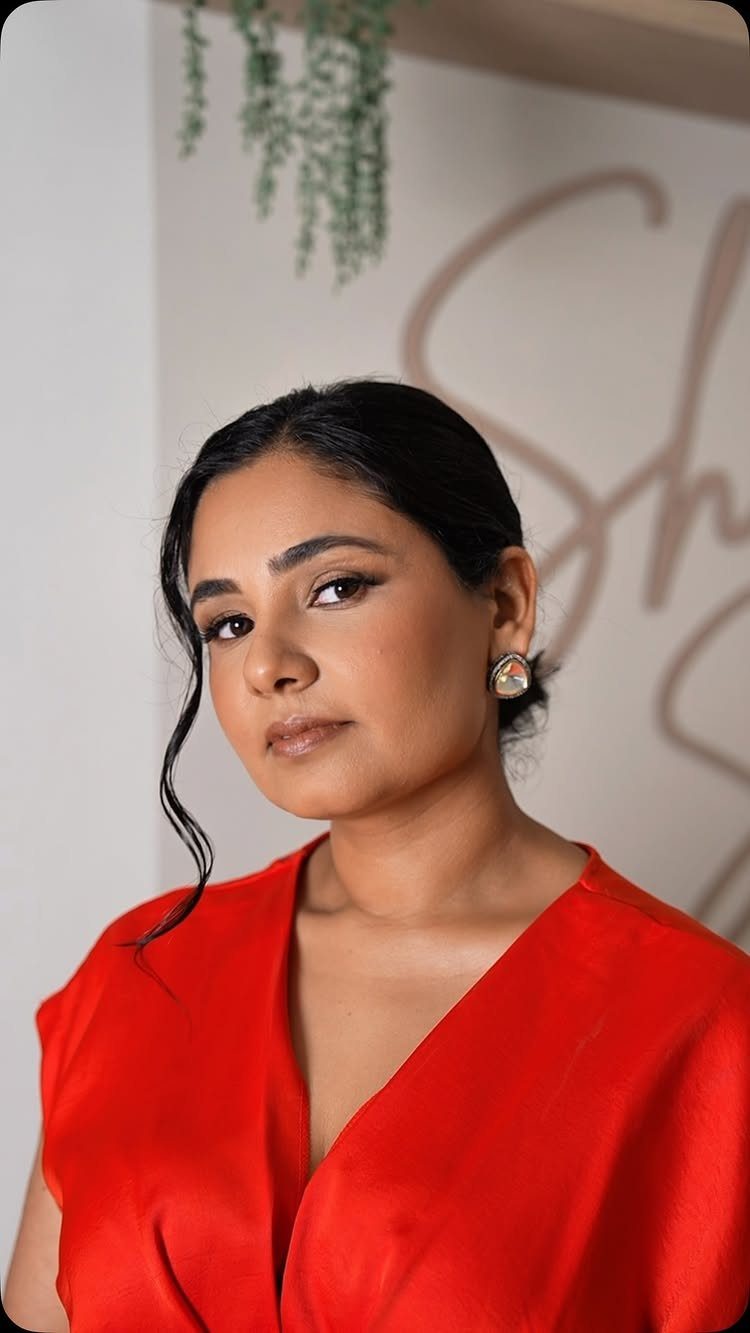 A woman with dark hair and neutral makeup wearing a red top and stud earrings.