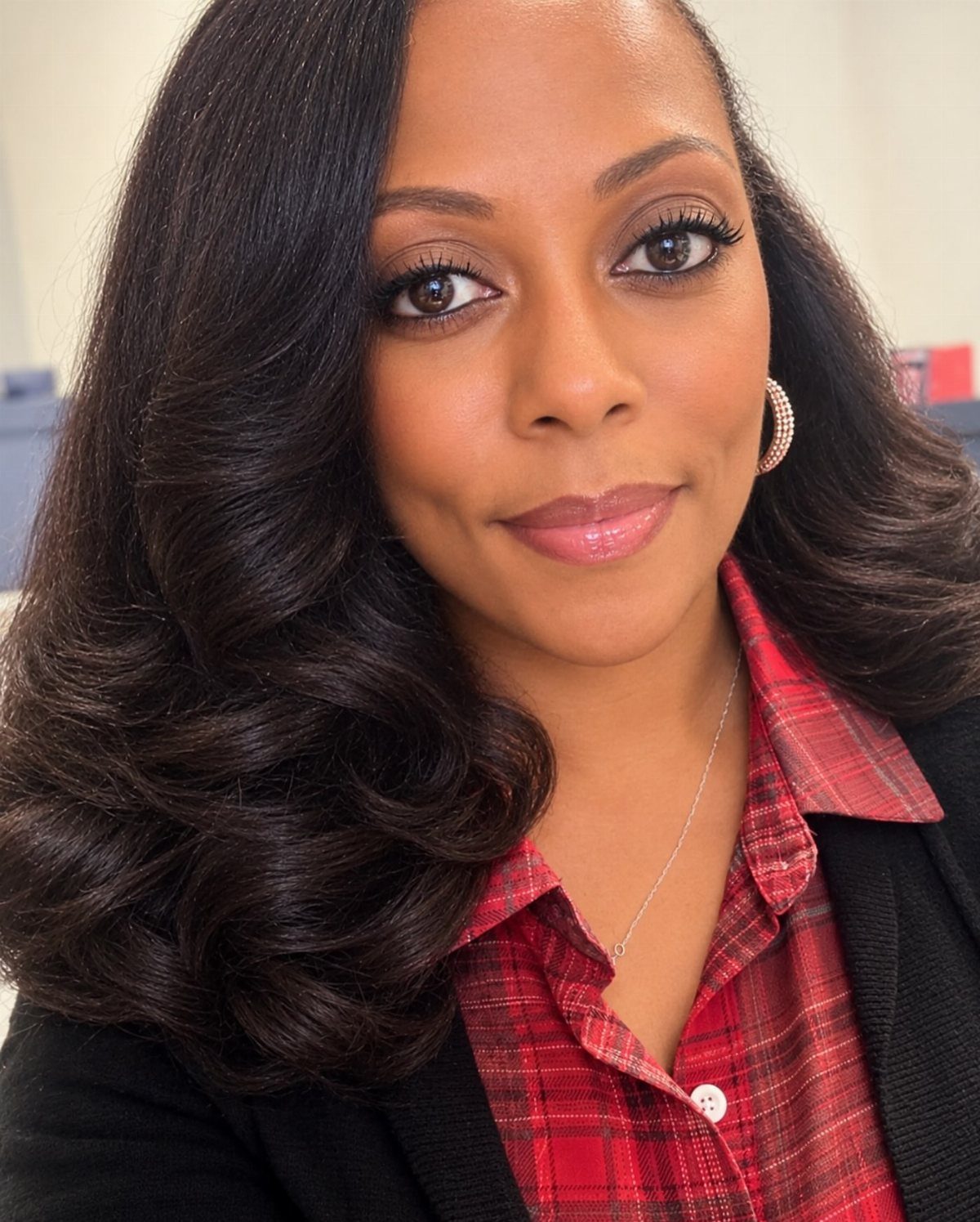 A smiling woman with dark wavy hair wearing a red plaid shirt and small hoop earrings.