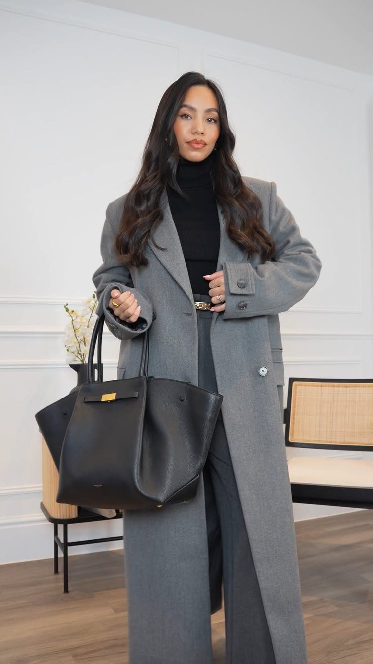 A woman wearing a grey overcoat, black turtleneck, and grey trousers holding a black tote bag.