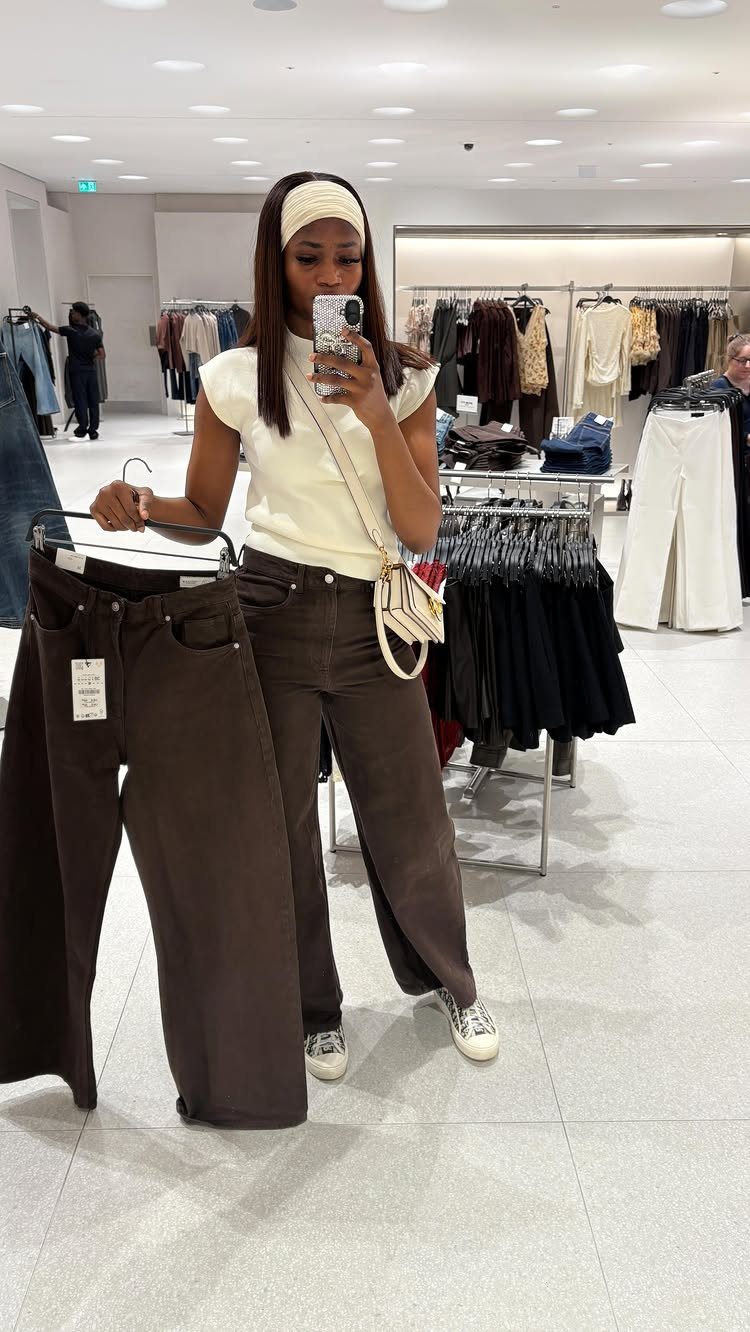 Woman holding up dark brown wide-leg pants while wearing a matching pair in a store.