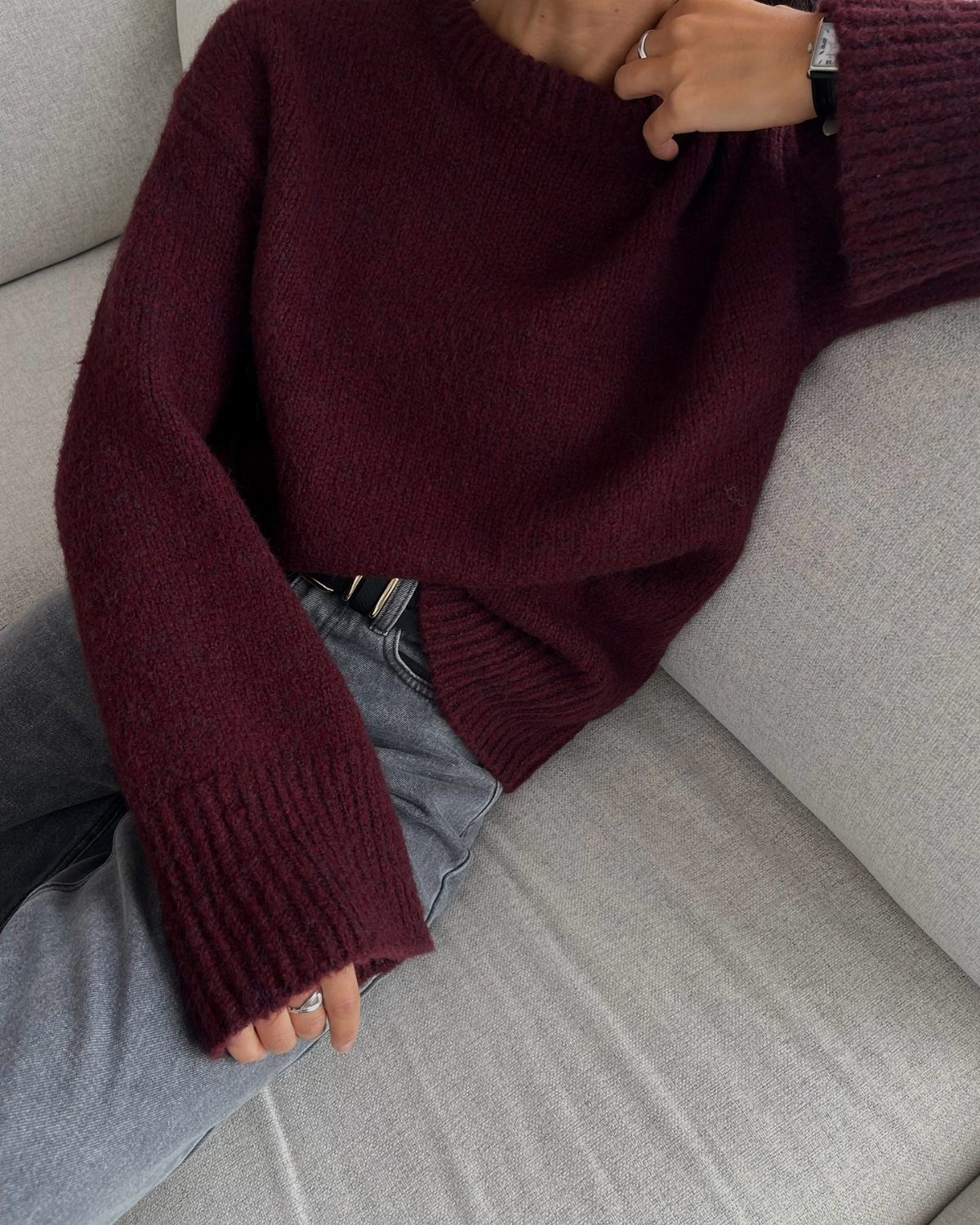 A person wearing a burgundy knit sweater and light wash blue jeans sitting on a sofa.