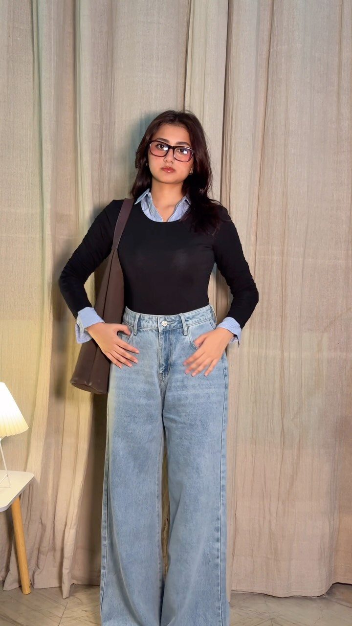 A woman posing in light wash wide-leg jeans and a black top against beige curtains.
