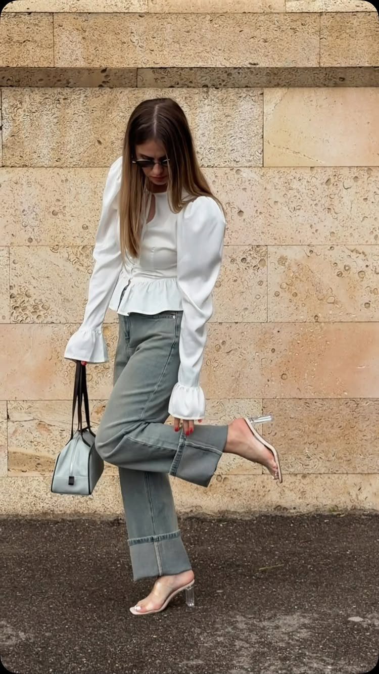 Woman posing in a white blouse, grey wide-leg jeans, and clear high-heeled sandals.