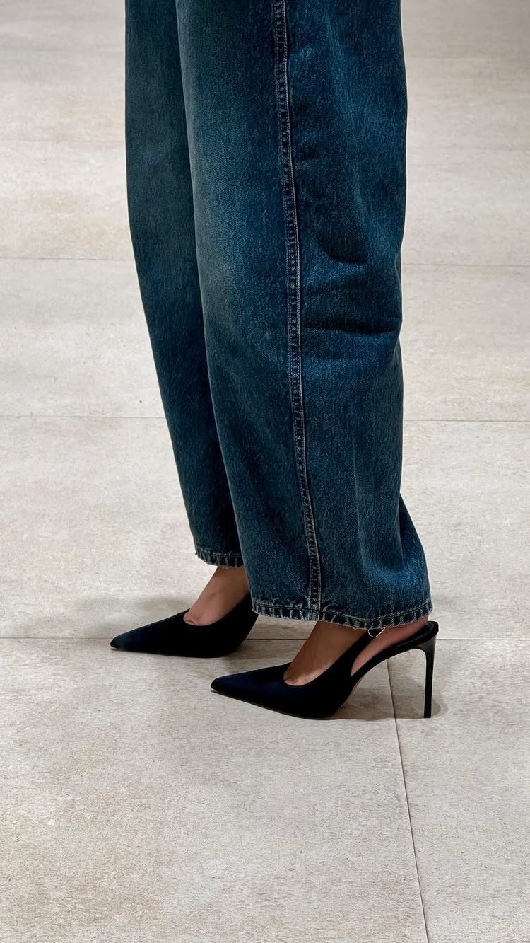 Person wearing wide-leg blue jeans and black pointed-toe high heels on a tiled floor.