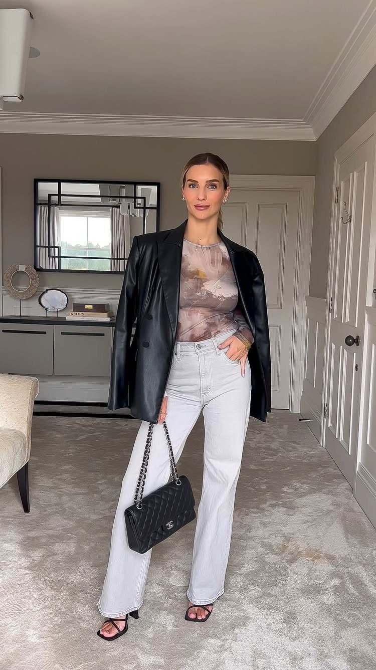 Woman posing in a black leather blazer, patterned top, light grey jeans, and black strappy heels.