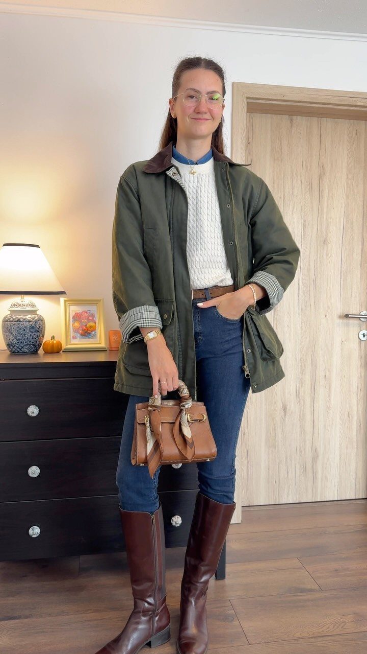 Woman wearing a white cable-knit sweater, blue jeans, and an olive green utility jacket.