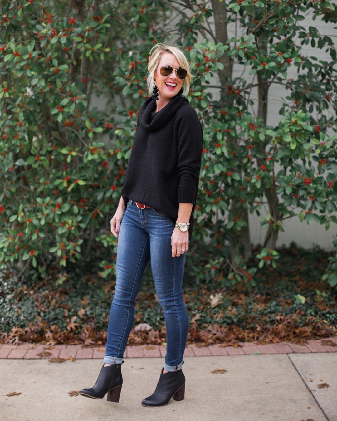 A woman wearing a black cowl neck sweater and blue skinny jeans standing outdoors.