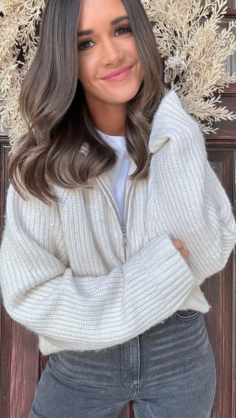 A smiling woman wearing a white ribbed half-zip sweater and black jeans outdoors.
