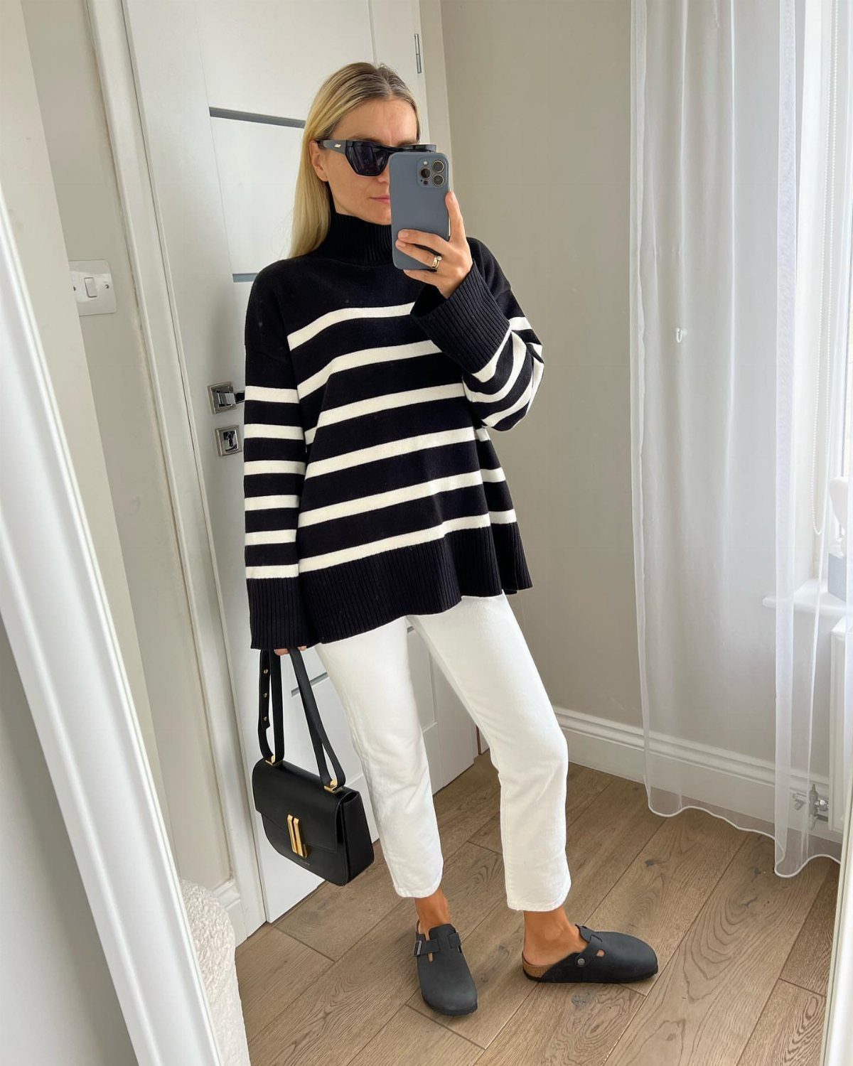 Woman in a striped oversized sweater and slim white jeans posing for a mirror selfie.
