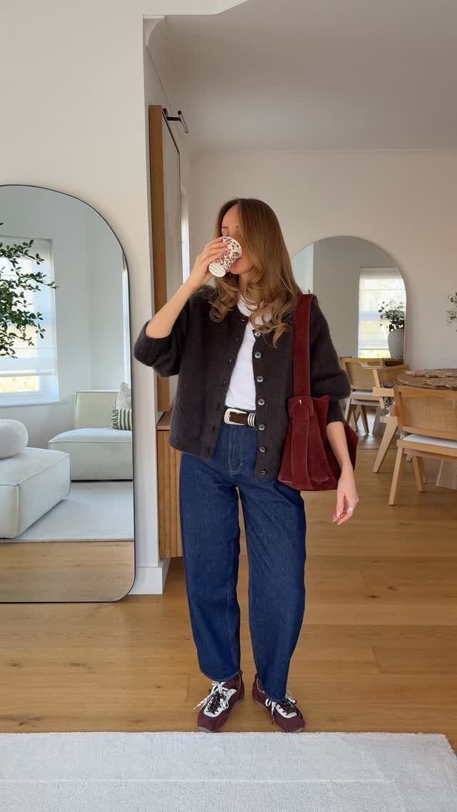 Woman wearing a dark oversized cardigan, white t-shirt, and wide-leg blue jeans standing indoors.