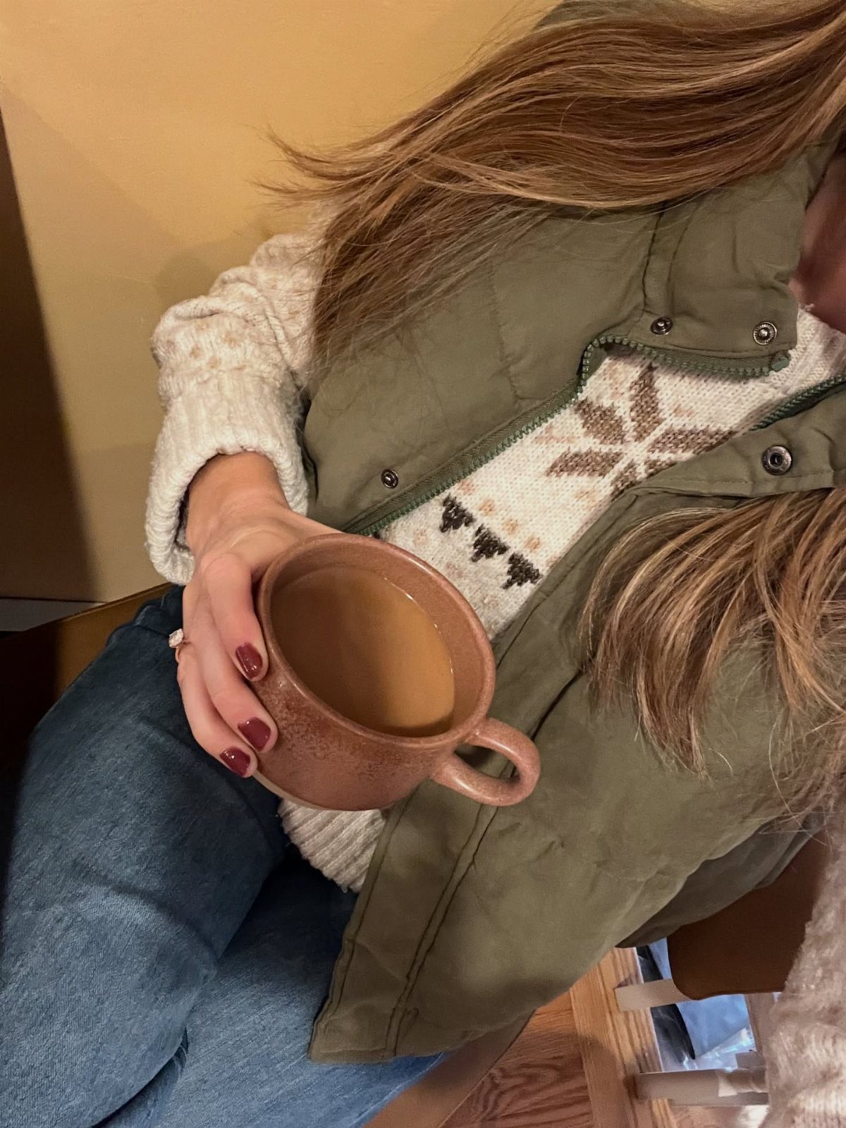 A person in a cream patterned sweater and green vest holding a coffee mug.