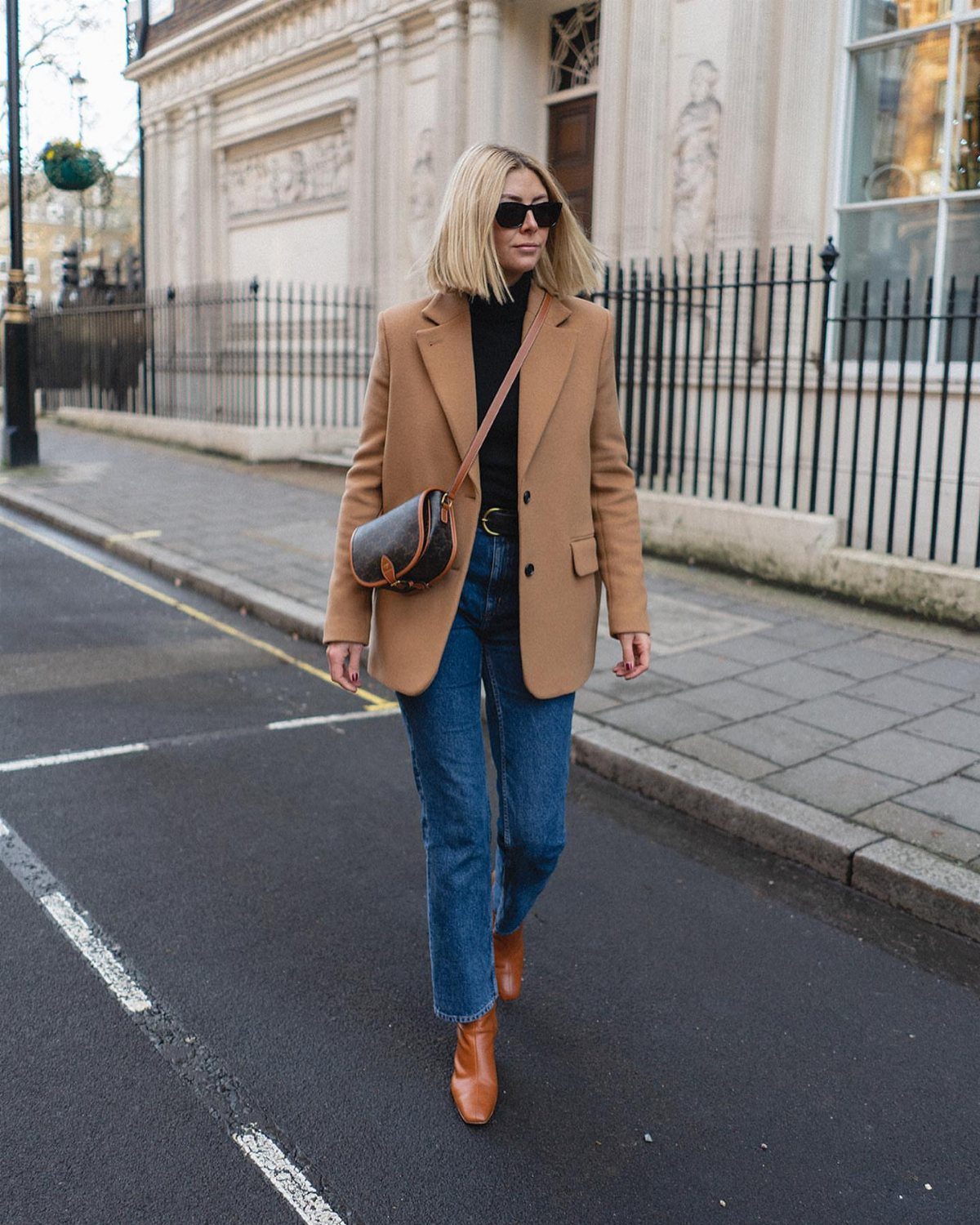 Blonde woman in a tan coat and blue jeans walking on a city street.
