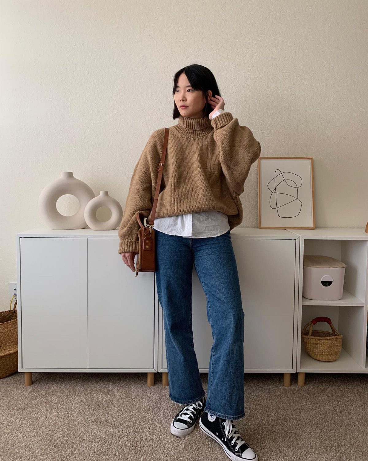 Woman wearing a brown turtleneck sweater, blue jeans, and black high-top sneakers standing indoors.