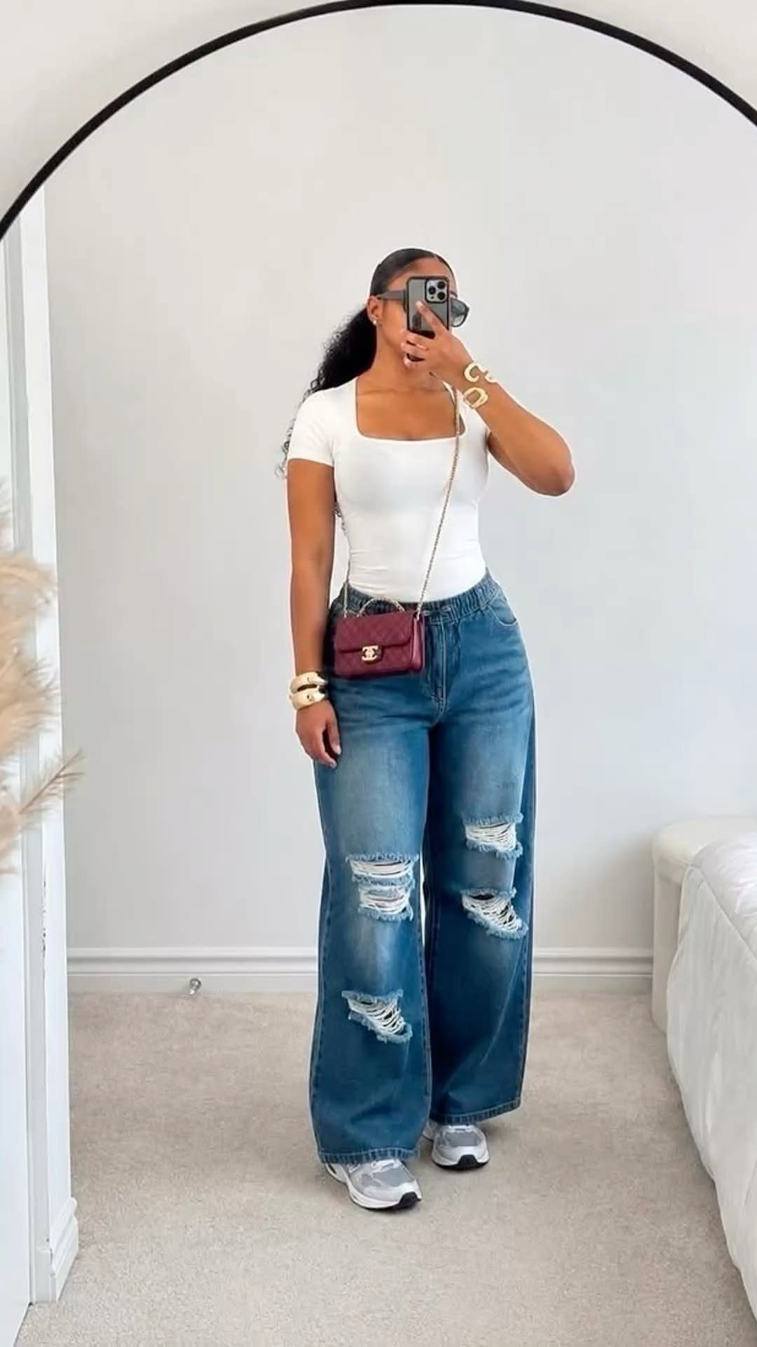 Woman in white tank top, distressed wide-leg jeans, and white sneakers posing for a mirror selfie.