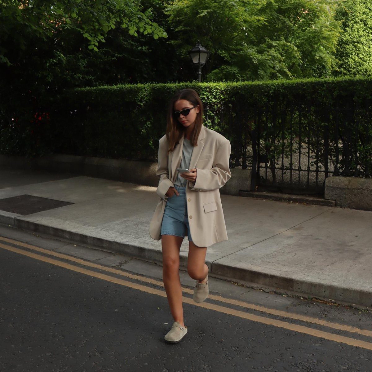 Woman walking on a city sidewalk wearing denim shorts, a beige blazer, and sunglasses.