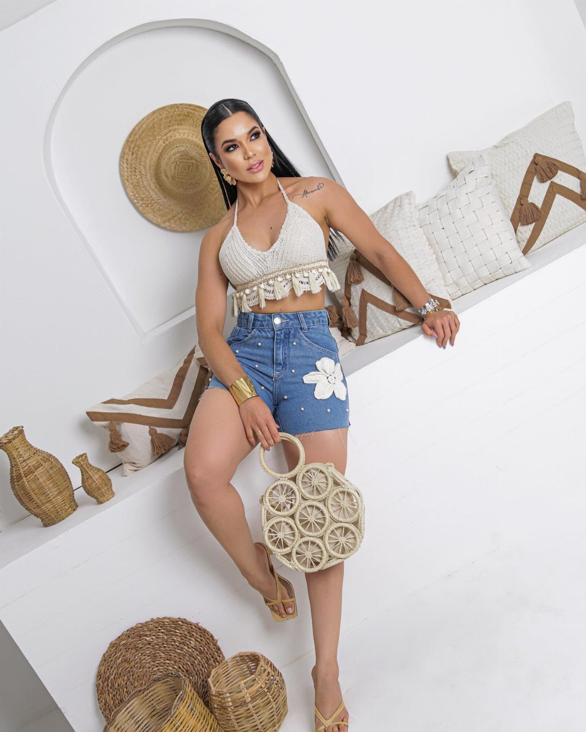 Woman in denim shorts and a white crochet halter top holding a woven bag.