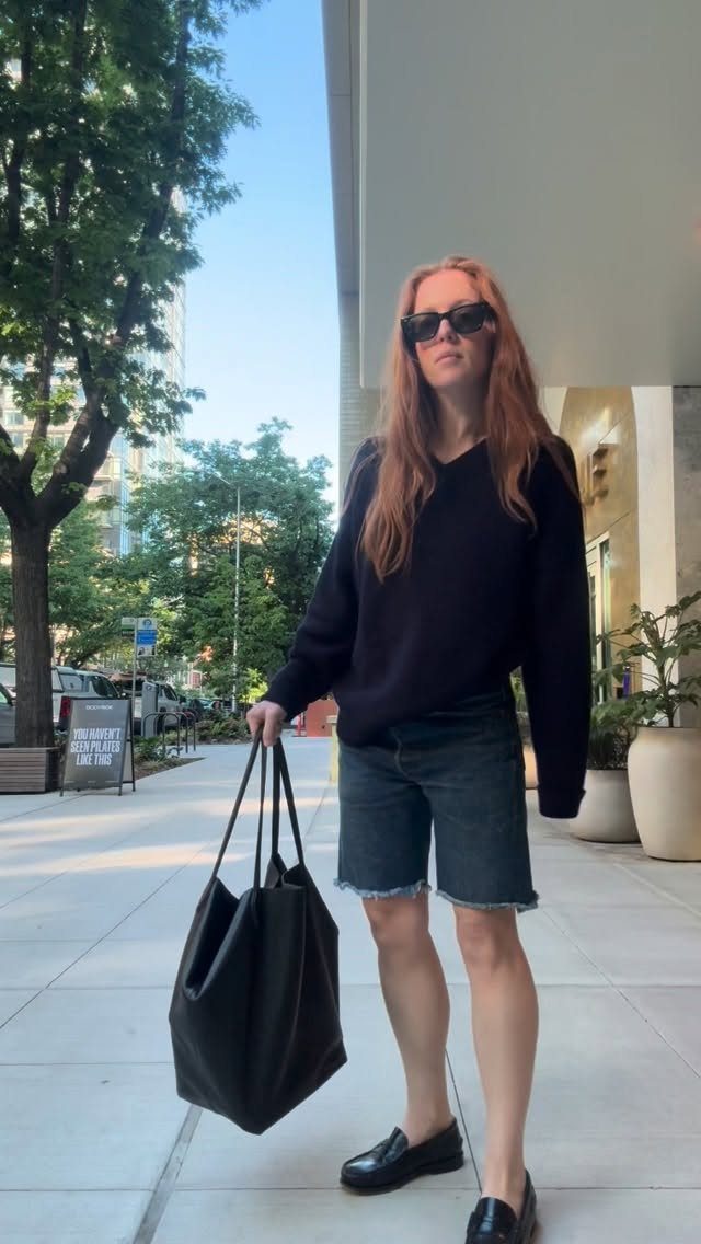 Woman wearing black sweater, denim shorts, and black loafers standing on a city sidewalk.