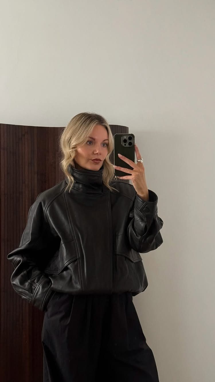Blonde woman wearing a black funnel-neck leather jacket and black trousers taking a mirror selfie.