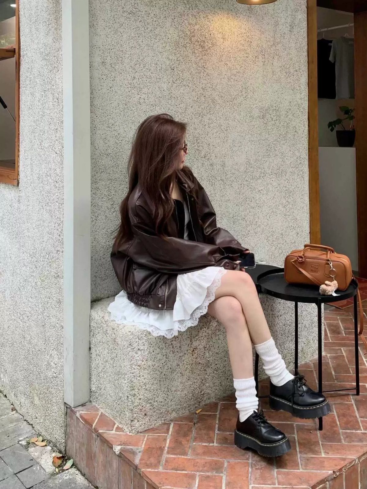 Woman sitting outdoors wearing a brown leather jacket over a white ruffled dress and black loafers.