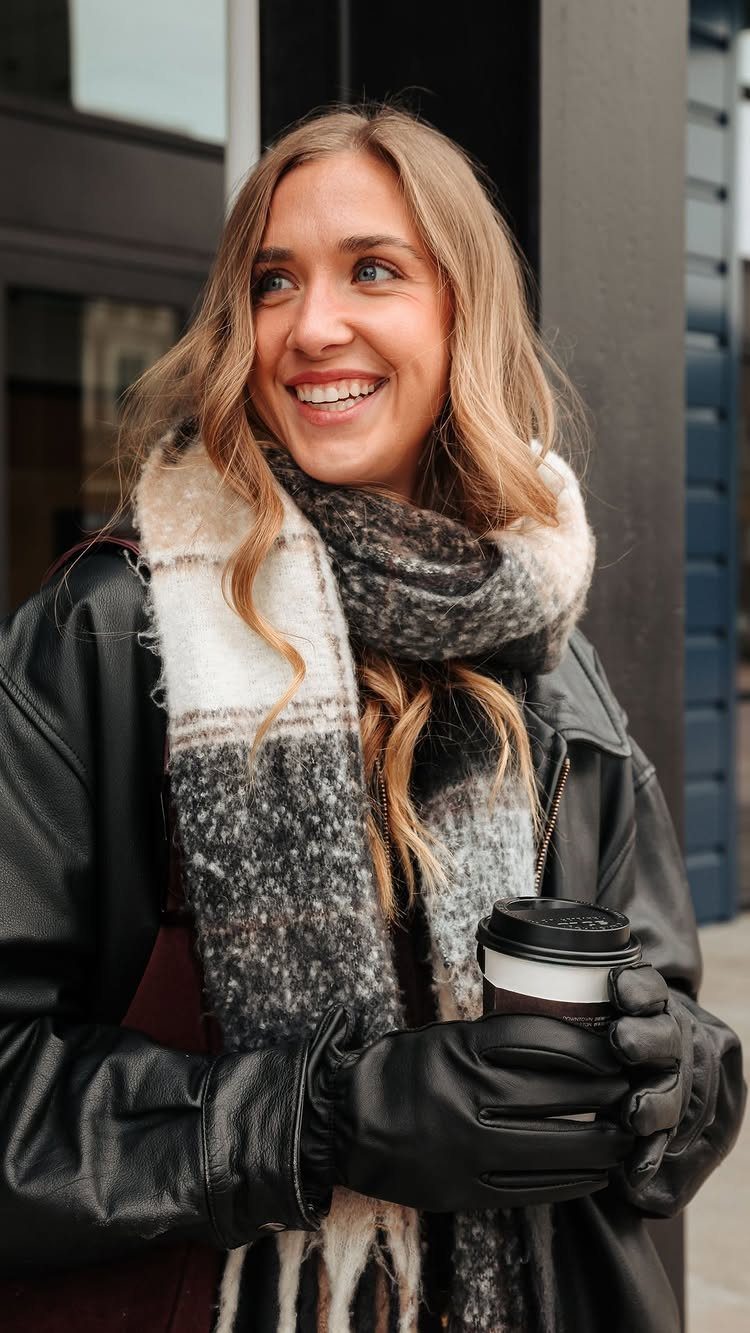 Smiling blonde woman wearing a black leather jacket and a thick patterned scarf while holding coffee.