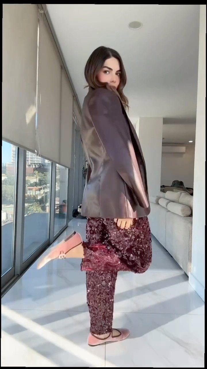 Woman posing in a brown leather blazer and sparkling burgundy sequined trousers indoors.