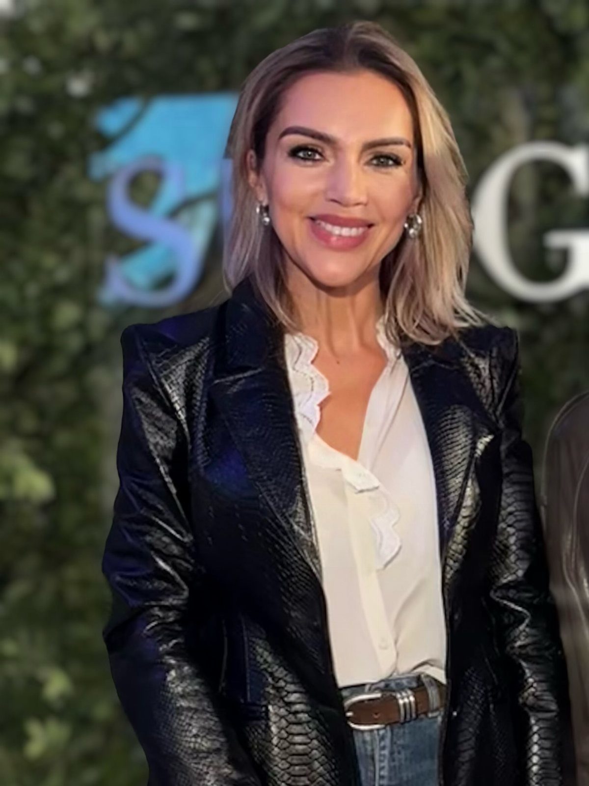A smiling woman wearing a black leather jacket over a white ruffled silk blouse.