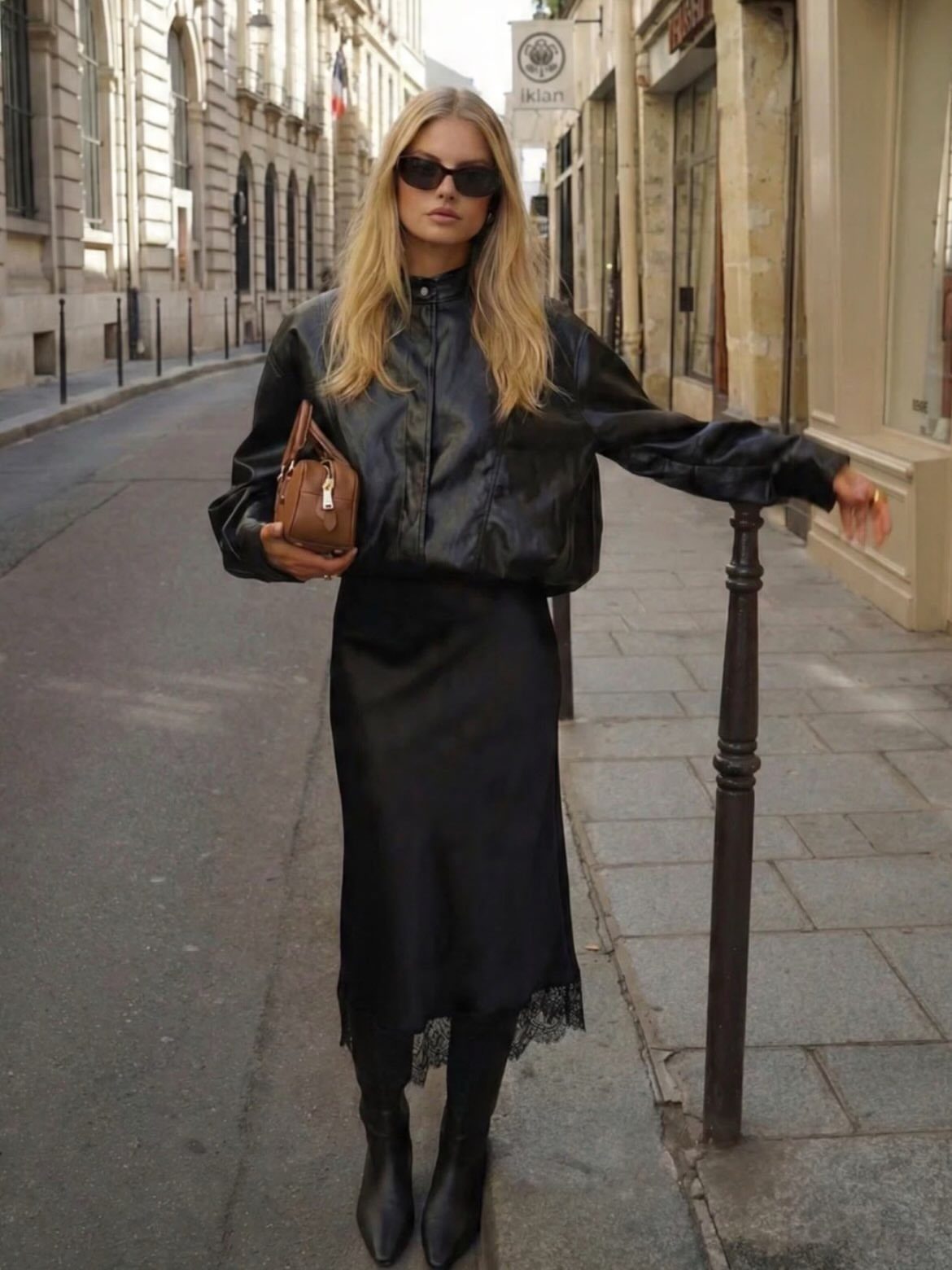 Blonde woman in a black leather jacket and a black satin slip skirt standing on a city street.