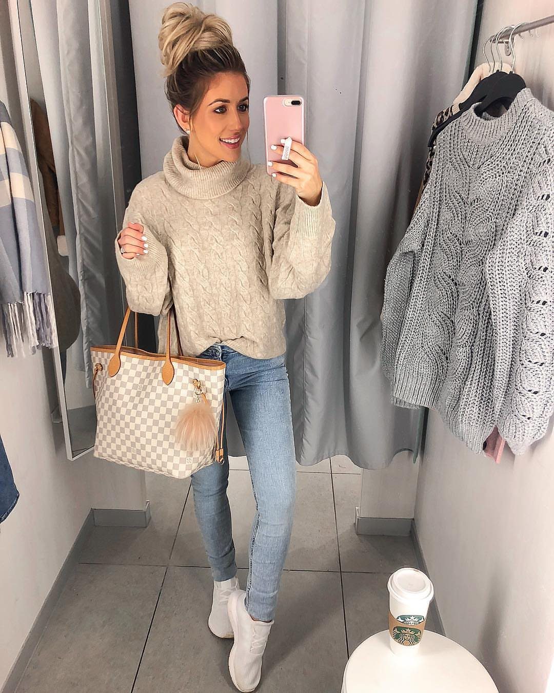 Woman in a beige knit sweater and light blue jeans posing for a mirror selfie.