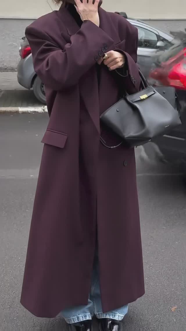 Person wearing a long plum coat and light blue jeans standing on a city street.